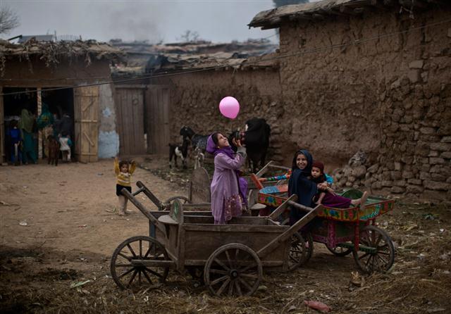 Một bé gái tị nạn người Afghanistan, ẵm đứa em trai trên tay, ngồi trên một chiếc xe kéo bằng gỗ xem đứa bạn đang chơi bong bóng, ở một khu dân cư nghèo thuộc ngoại ô Islamabad, Pakistan